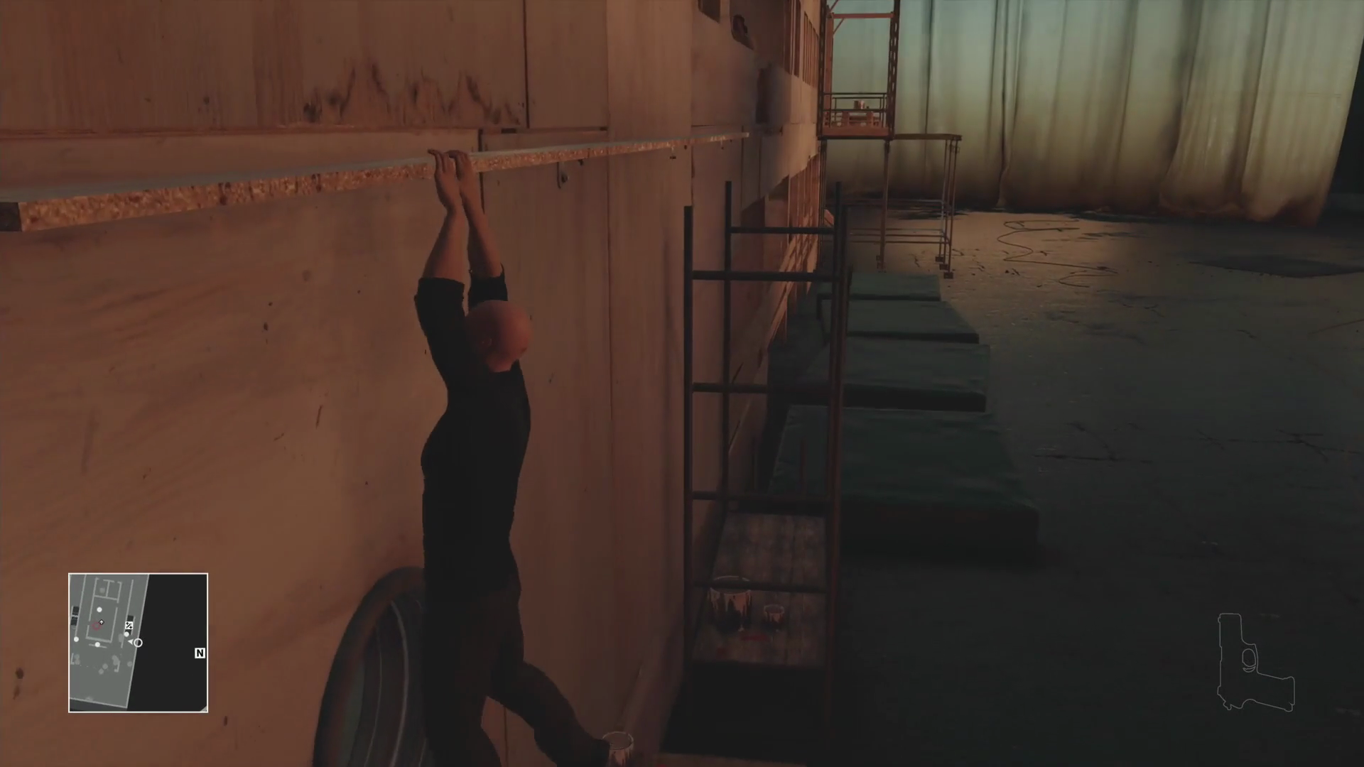 Screenshot aus HITMAN: Agent 47 hangelt an der Seitenwand einer aus Spanplatten gebauten Yacht, auf dem Boden liegen dicke Sportmattten.