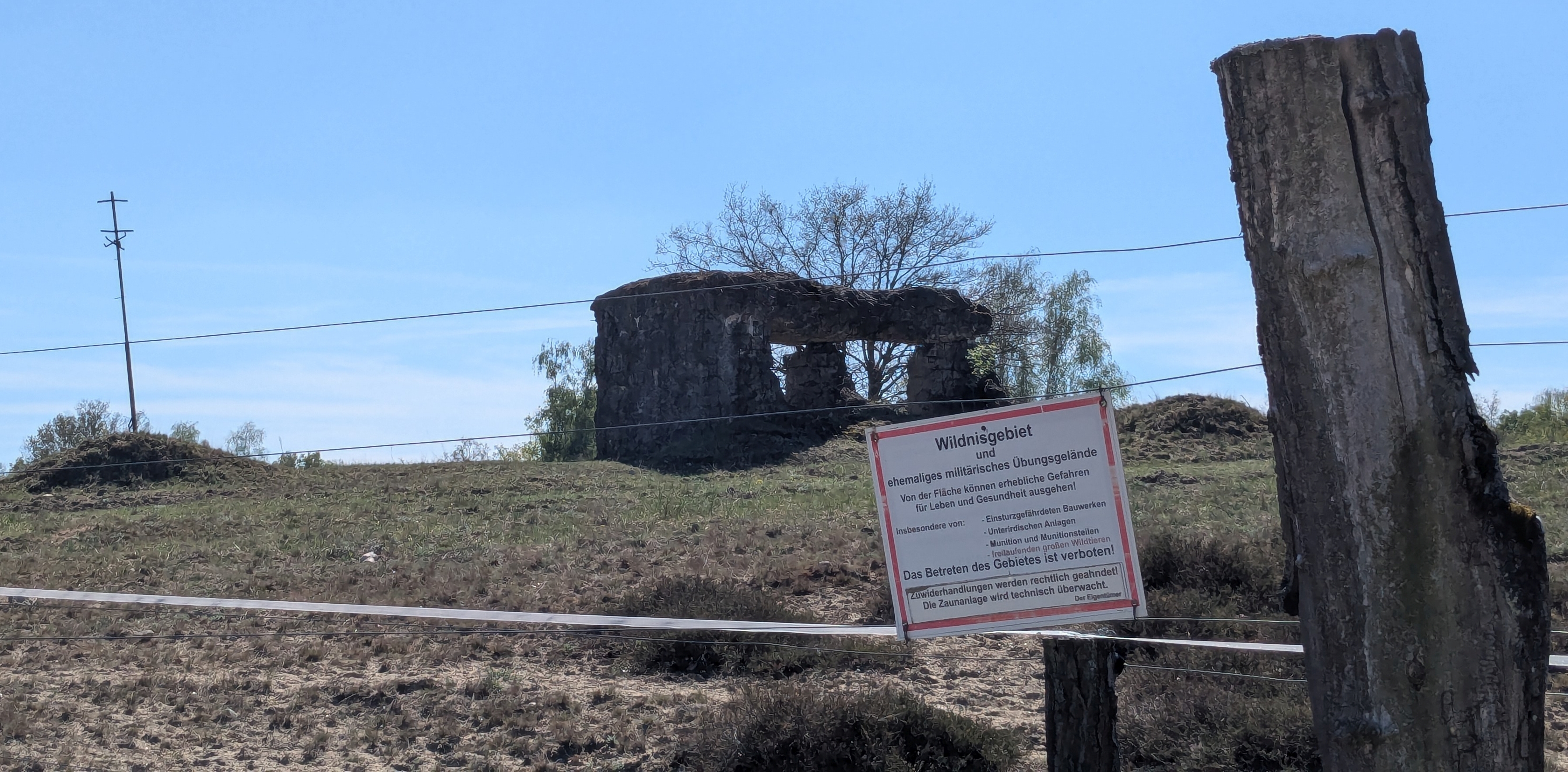 Auf einem weitläufigen, kargen Gelände steht die Ruine eines alten Betonbaus, der sichtlich von Zeit und Witterung gezeichnet ist. Die Natur hat das Areal teilweise zurückerobert – Bäume wachsen aus und um die zerfallenen Mauern. Im Vordergrund weist ein deutlich sichtbares Warnschild darauf hin, dass es sich um ein ehemaliges militärisches Übungsgelände handelt. Es warnt vor Gefahren wie einsturzgefährdeten Bauwerken oder Munitionsresten und macht klar, dass das Betreten streng verboten ist. Spanndraht und ein abgesägter Baumstamm markieren die Grenze des gesperrten Gebietes.