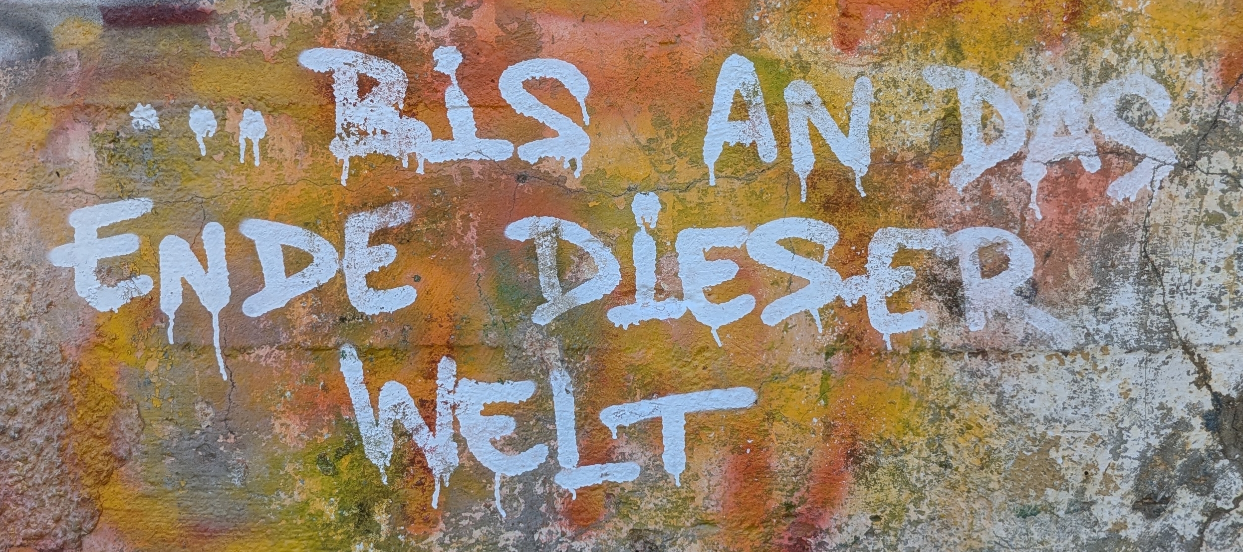 Das Bild zeigt eine bunt besprühte Wand, deren Oberfläche vom Alter bröckelt und von mehreren abgenutzten Farbschichten auf rissigem Beton geprägt ist. In kräftigem Weiß leuchtet der Text „…BIS AN DAS ENDE DIESER WELT“ auf orange-rotem Untergrund.
